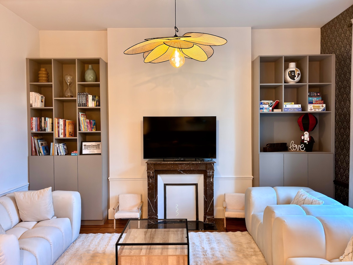 The living area features light-colored sofas arranged around a central glass-topped coffee table. Dark shelves hold various books and decorative items, while a large television is mounted above a stylish marble fireplace. A unique yellow light fixture adds a distinct touch to the space.