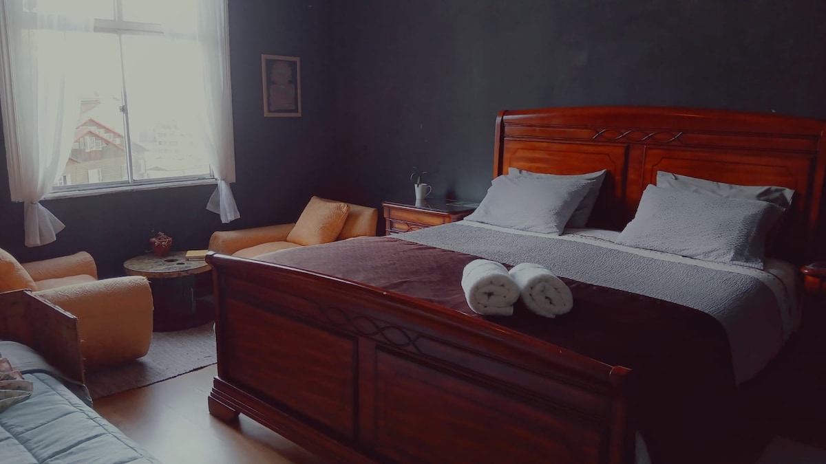 A spacious room features a large wooden bed with neatly arranged linens. Two plush armchairs are placed near a small side table, offering a cozy seating area. Natural light enters through a large window, illuminating the dark walls and enhancing the room's inviting feel.