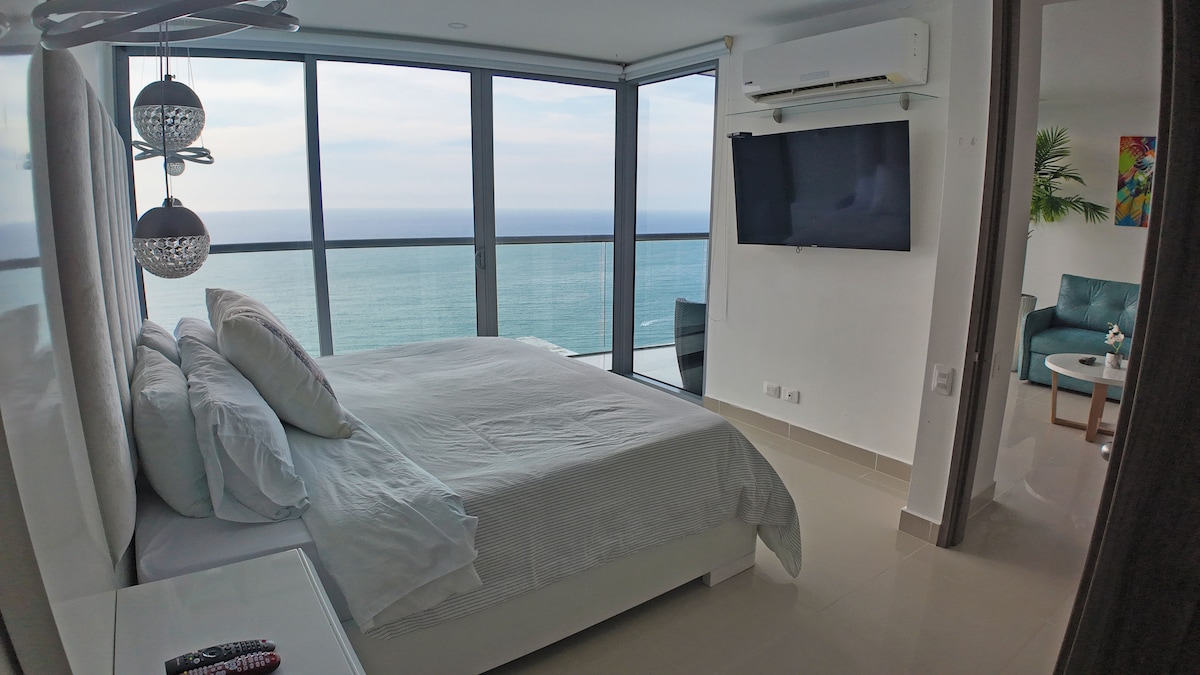 A modern bedroom features a spacious bed with crisp white linens, positioned near floor-to-ceiling windows that offer panoramic views of the ocean. A flat-screen TV is mounted on the wall, and a comfortable seating area is visible in the corner.
