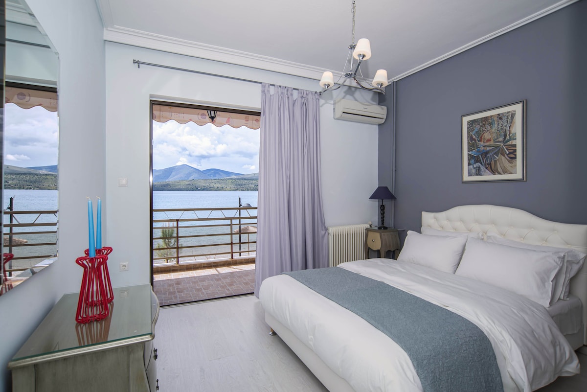 A serene bedroom features a spacious bed with crisp white linens and a soft blue throw blanket. Large glass doors open to a balcony that overlooks the sea, allowing natural light to fill the room. A decorative mirror and a lamp beside the bed add to the inviting decor.