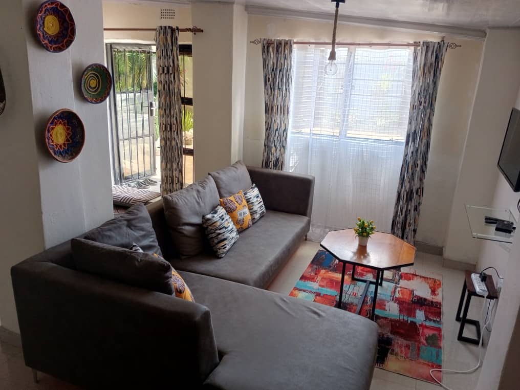 A spacious living area features a gray sectional sofa adorned with colorful cushions. A wooden coffee table rests on a vibrant area rug, while large windows allow natural light to brighten the room. Decorative wall plates add a touch of character to the space.