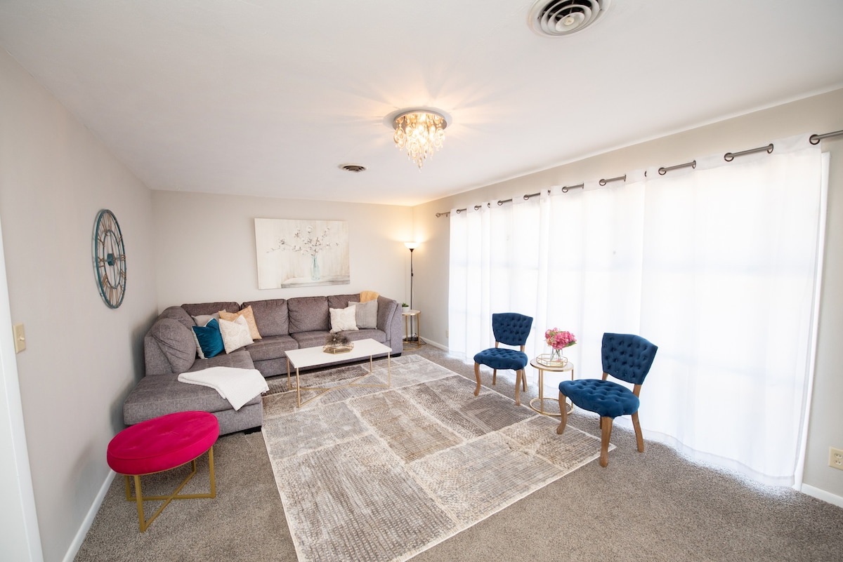 A bright living area is presented, featuring a spacious sectional sofa and a central coffee table. Natural light filters through sheer curtains, illuminating a plush area rug. Two elegant chairs with blue upholstery and a round pink table add a touch of color to the neutral decor.