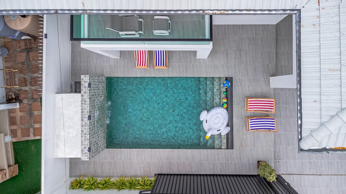 An aerial view showcases a serene private pool with crystal-clear water, surrounded by stylish lounge chairs with colorful striped fabric. An inflatable float adds a playful touch, while well-maintained greenery lines the pool area, enhancing the inviting outdoor space.