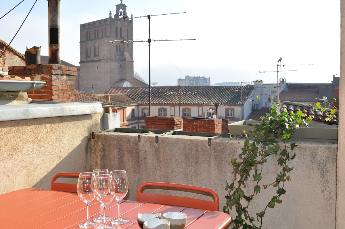 A rooftop terrace is shown, featuring a red dining table set with four clear drinking glasses. The view includes rooftops of nearby buildings and the prominent bell tower of a church in the background, providing an inviting space for outdoor meals.