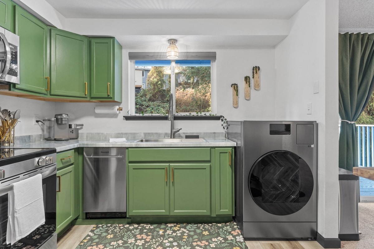 A fully equipped kitchen showcases vibrant green cabinetry and modern appliances, including a stove, dishwasher, and washing machine. Natural light floods the space through a window, highlighting a stylish countertop. A patterned rug adds warmth to the room, enhancing the overall functionality.