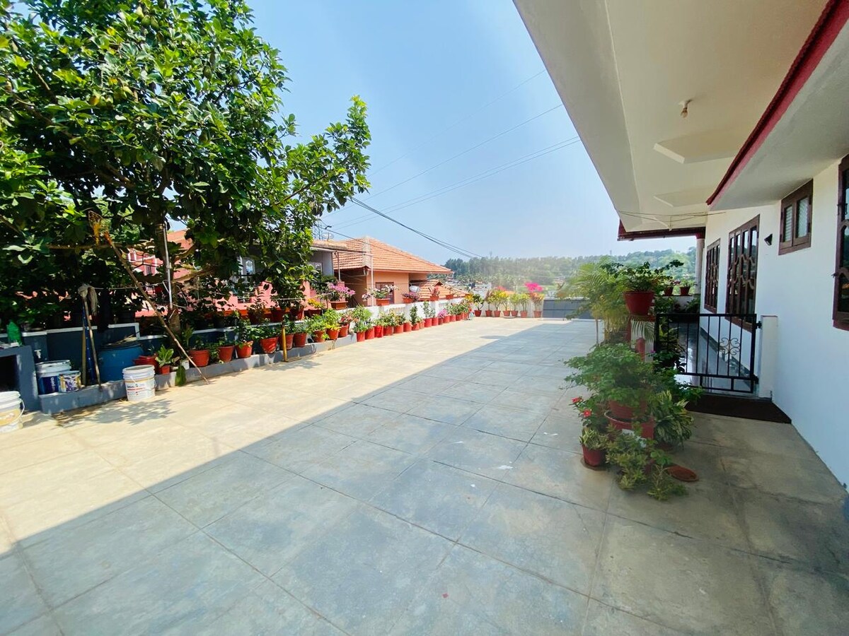 An expansive paved area is surrounded by a vibrant garden filled with potted plants. The space features a mix of green shrubs and flowering plants, while a staircase leads to a covered entrance. Clear skies create a bright and inviting atmosphere.