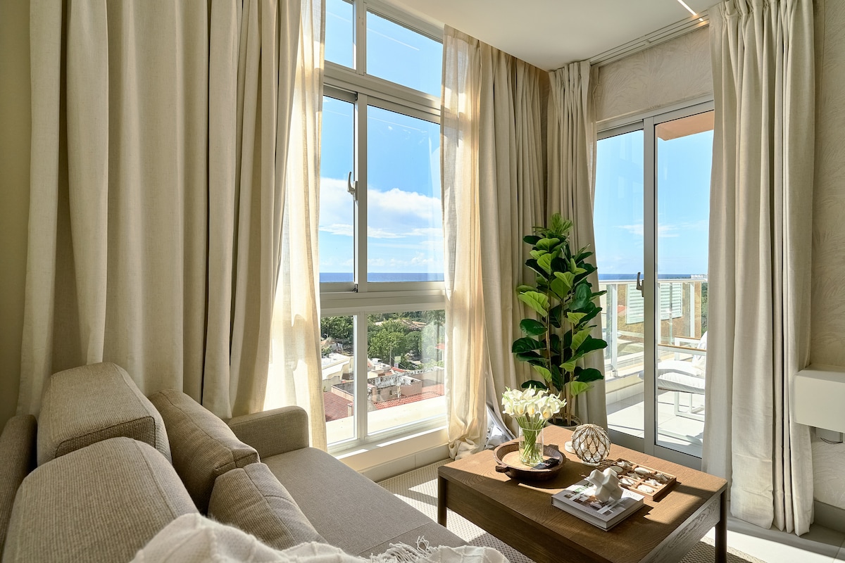 A welcoming living area is illuminated by natural light from large windows. Soft, neutral curtains frame the view, while a plush sofa invites relaxation. A coffee table holds decorative items and a small flowering plant, enhancing the comfort of the space.