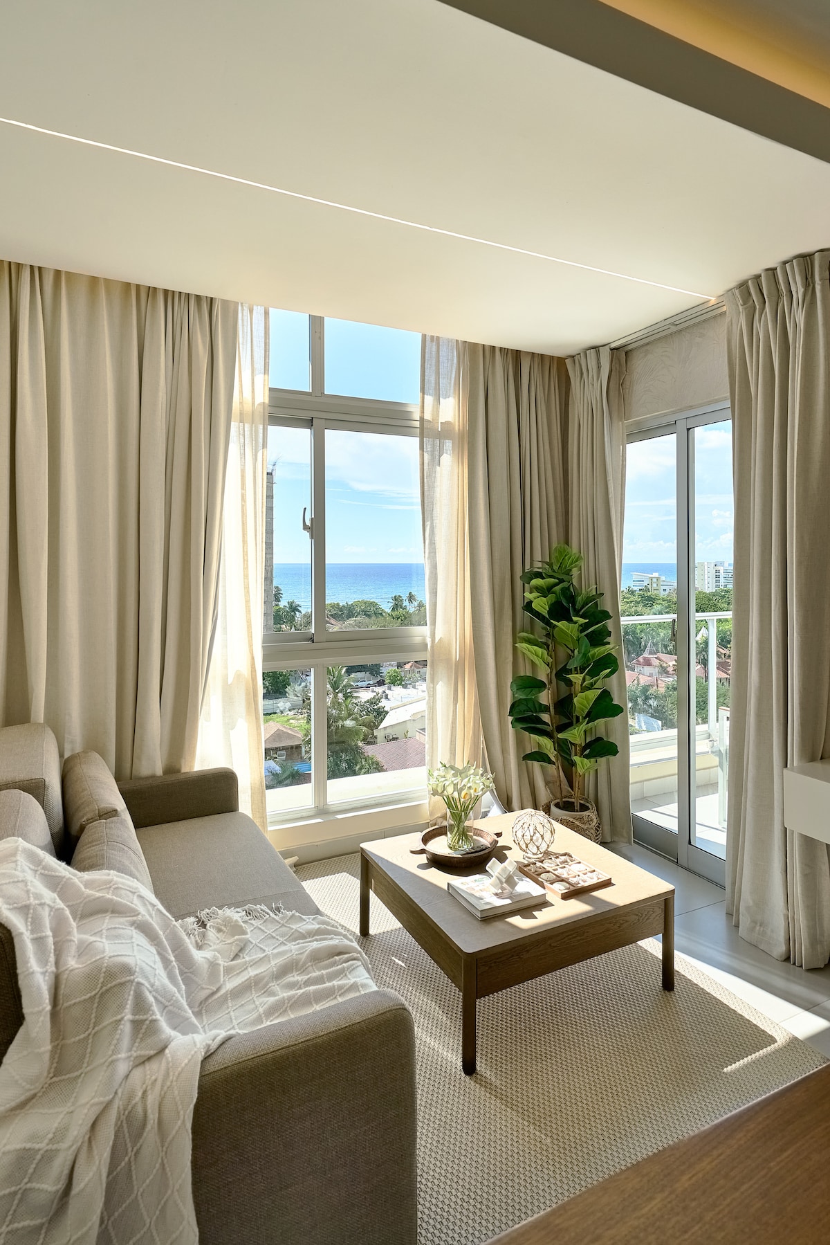 A bright living area is illuminated by natural light streaming through large windows, offering views of the coastline. A neutral-toned sofa is positioned beside a small coffee table, adorned with decorative items. Light-colored curtains frame the windows, enhancing the airy ambiance.