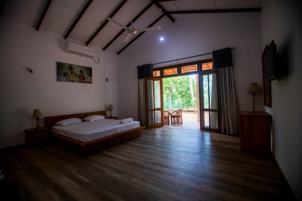 A spacious room features a large bed with a simple wooden frame, situated under a high, sloped ceiling. Two doors open to a serene view of the outside, while natural light fills the space, complemented by curtains and wooden furniture.