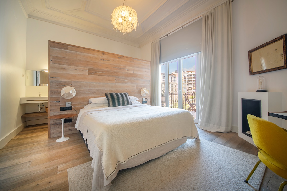 A spacious bedroom features a wooden accent wall and a large bed dressed in neutral tones. Natural light enters through a wide window, complemented by soft curtains. A corner workspace with a chair and modern light fixtures adds functionality to the inviting setting.