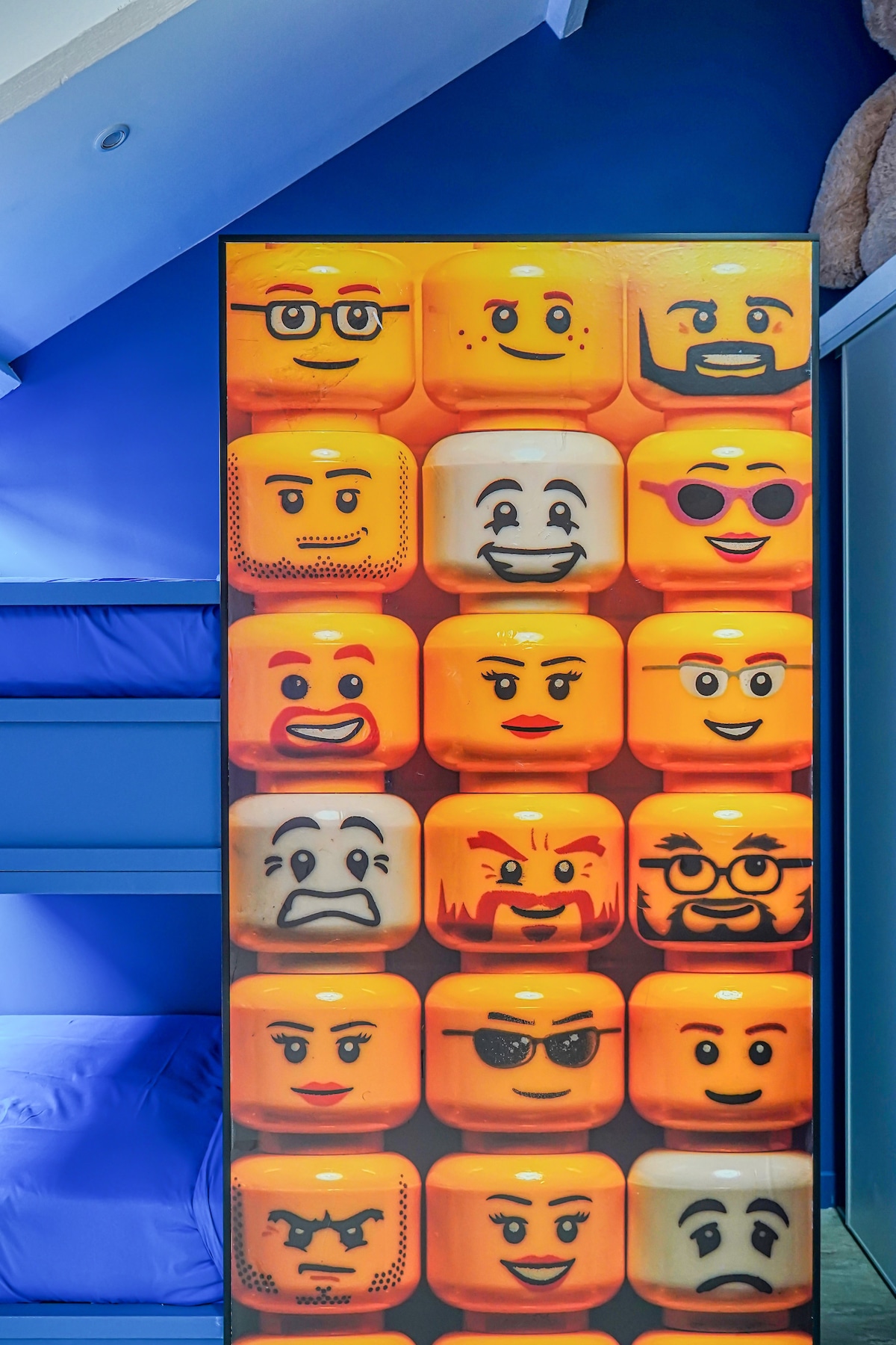 A vibrant wall display showcases various LEGO character faces, each capturing unique expressions against a bright orange background. The characters' playful appearances enhance the fun atmosphere of the loft, contributing to its immersive LEGO theme.