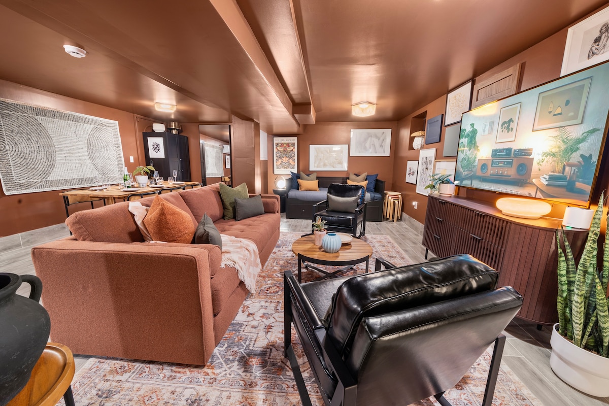 The entertainment lounge features a warm color palette with rich brown walls and stylish furnishings. A large, comfortable sofa and two armchairs invite relaxation. Art pieces adorn the walls, and a flat-screen TV is positioned for viewing. Soft lighting creates a cozy atmosphere throughout the space.