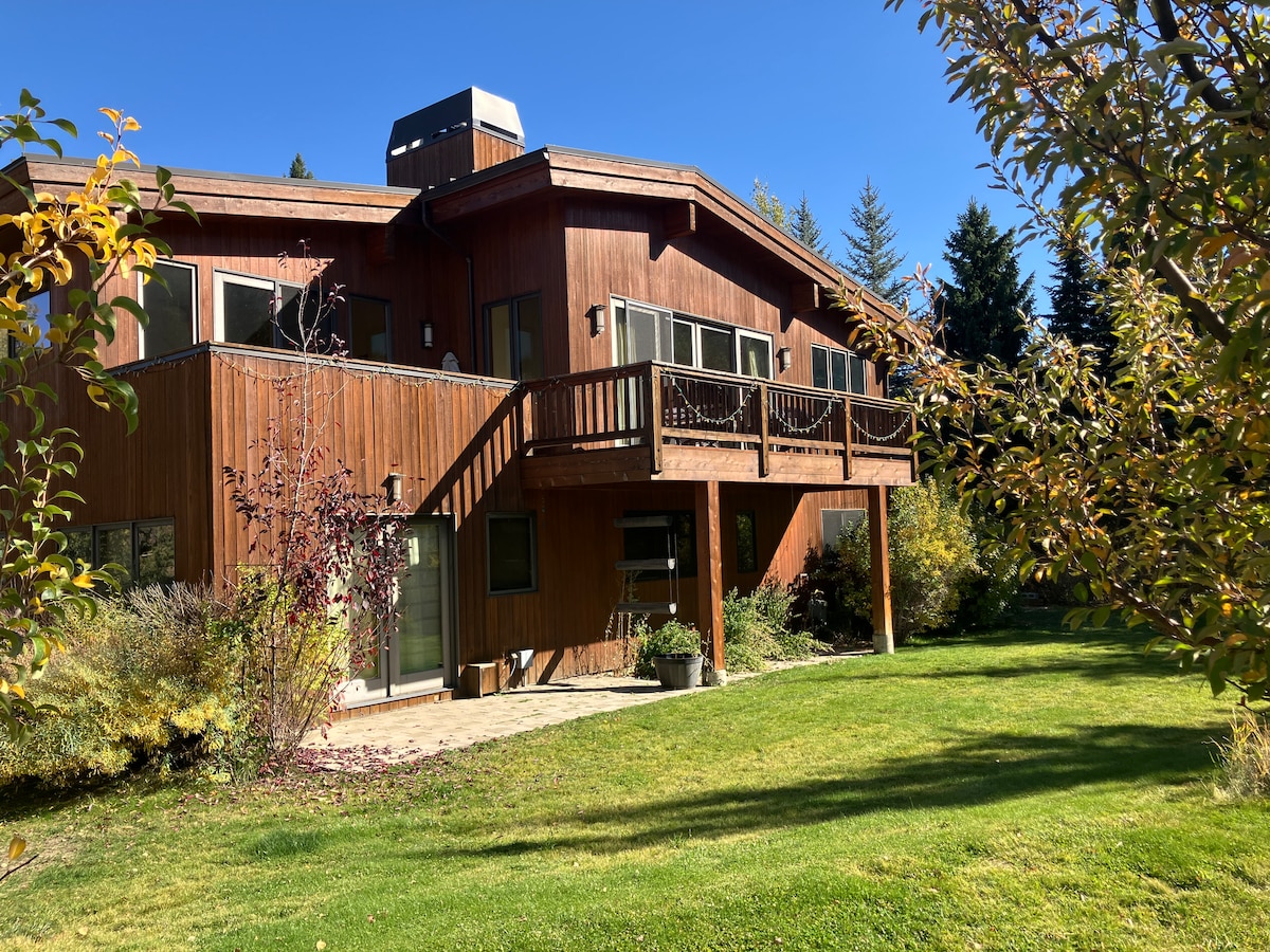 The exterior of the chalet is presented, showcasing a wooden façade with multiple windows and a large deck. Lush greenery and flowering plants frame the space, complementing the natural setting. A stone patio area is visible in the foreground, inviting outdoor relaxation.