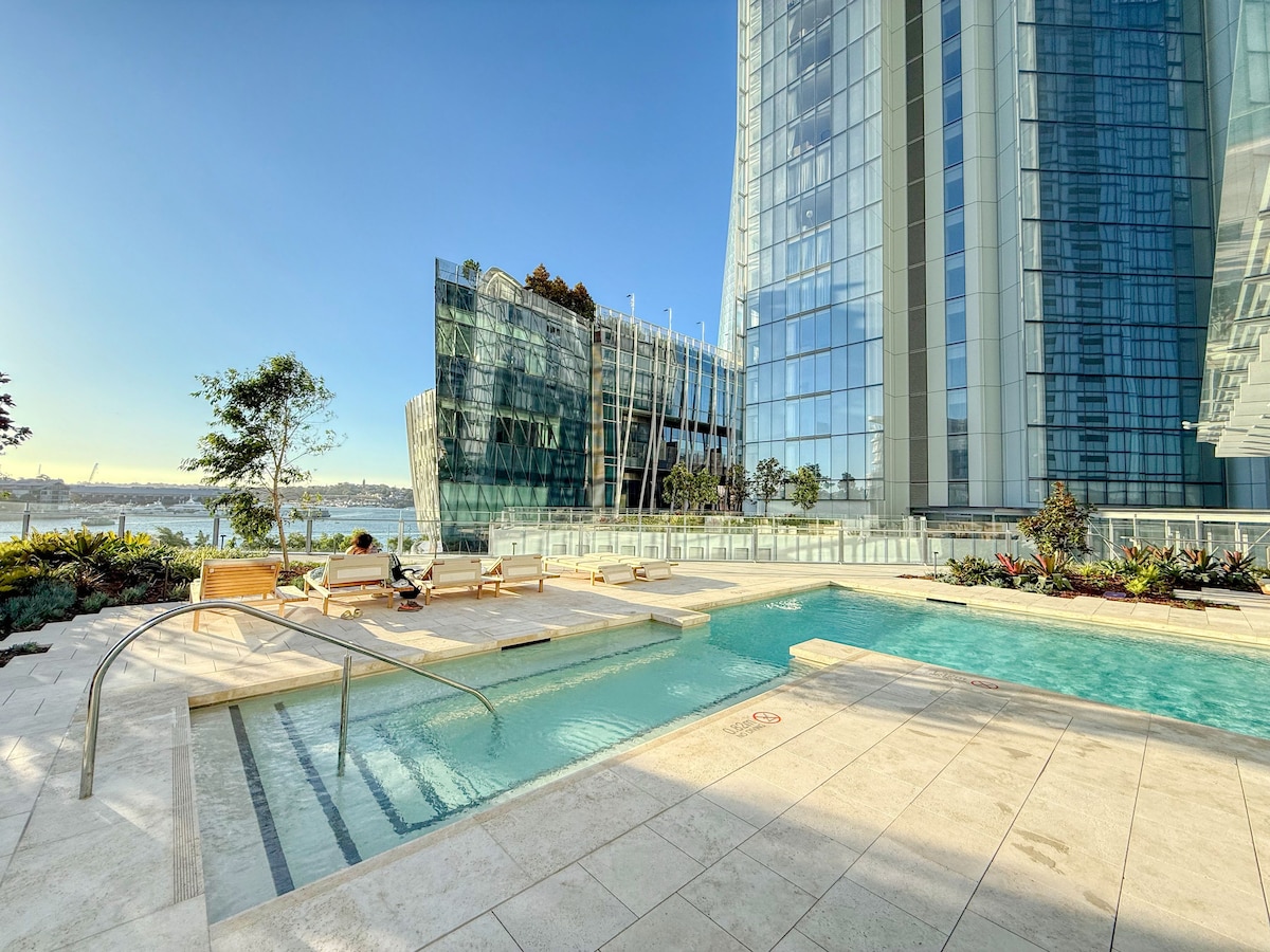 An outdoor swimming pool area is highlighted by smooth stone tiles and modern landscaping. Clear waters reflect the sky, while comfortable wooden loungers are positioned beside the pool, inviting relaxation. Surrounding greenery adds a natural touch to the luxurious environment.