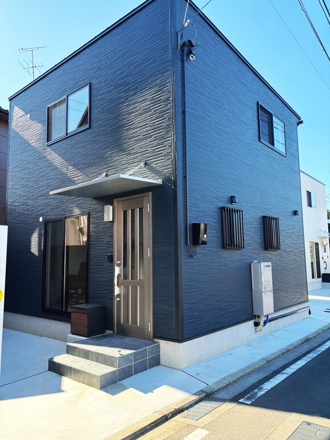 The exterior of the home features a simple, stylish design with dark panels and large windows. A small flight of steps leads to the entrance, while external wall accents provide visual interest. The setting is in a quiet residential area.