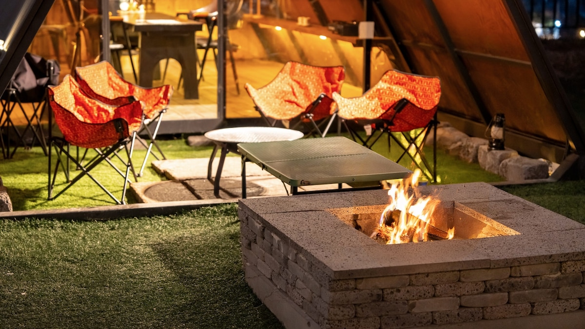 An outdoor area features a fire pit surrounded by comfortable seating. Orange-patterned chairs face the fire, while a low table sits nearby. The space is partially illuminated by the warm glow of the flames, creating a cozy ambiance in the setup.