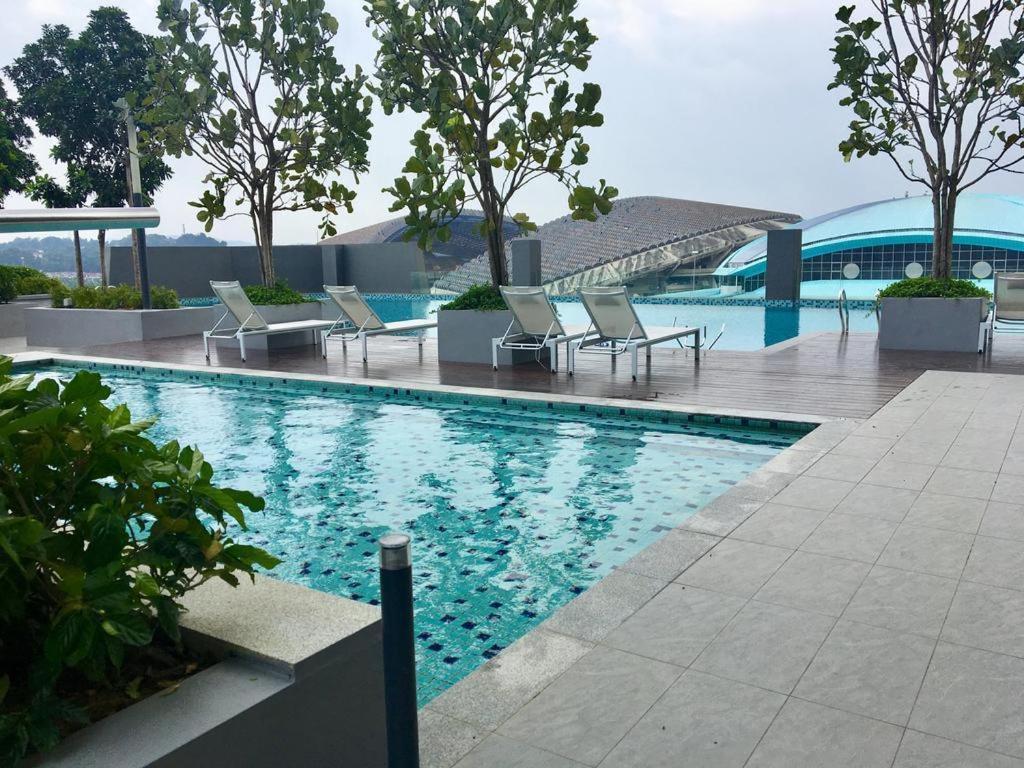 A serene pool area is surrounded by greenery, featuring several lounge chairs positioned along the water's edge. The pool's clear surface reflects the sky, while large trees provide a sense of tranquility. In the background, a modern structure is visible, enhancing the urban atmosphere.