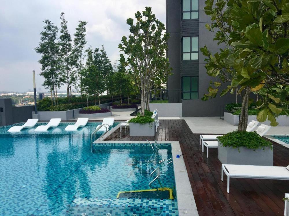 A serene pool area is featured with clear blue water reflecting the sky. White lounge chairs are positioned beside the pool, surrounded by landscaped greenery. Modern planters are placed along the edge, enhancing the tranquil setting.