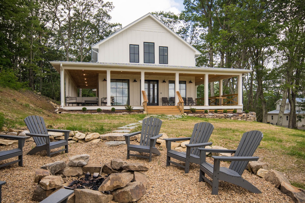The modern farmhouse is shown from the exterior, featuring a wrap-around porch with multiple seating areas. In the foreground, eight Adirondack chairs surround a fire pit, with a gravel path leading to the entrance. Lush greenery and trees create a tranquil outdoor setting.