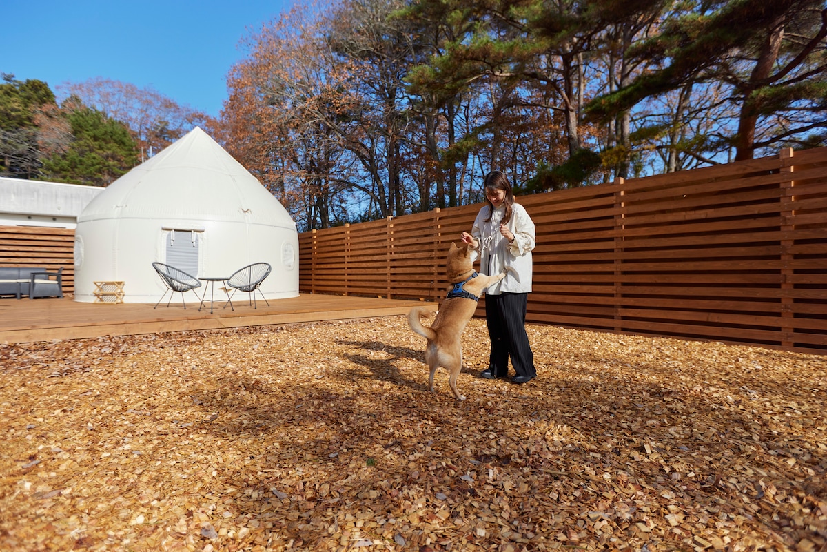 A spacious outdoor area features a textured ground of wood chips, surrounded by wooden fencing for privacy. A dome-shaped structure is visible beside the decking, along with a small table and two chairs. A person interacts with a dog, fostering a cozy atmosphere.