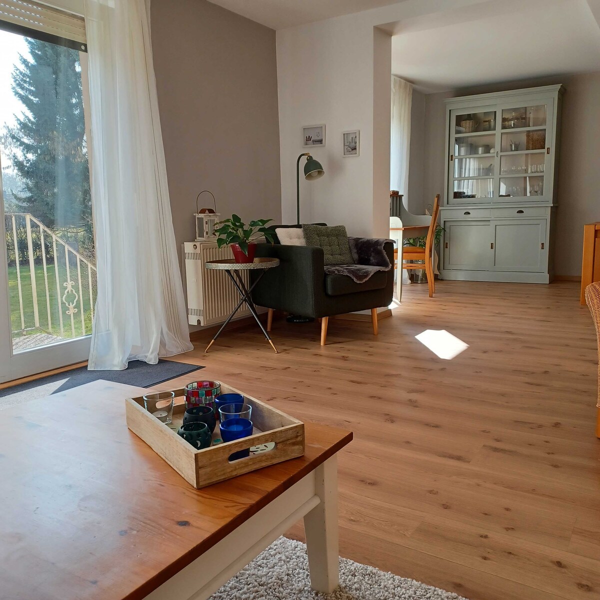 A cozy living area is highlighted by a relaxing seating arrangement, featuring a green armchair and a wooden table. Sunlight streams in through large glass doors, illuminating the natural wood flooring. A sideboard displays decorative items, with a plant adding a touch of greenery.