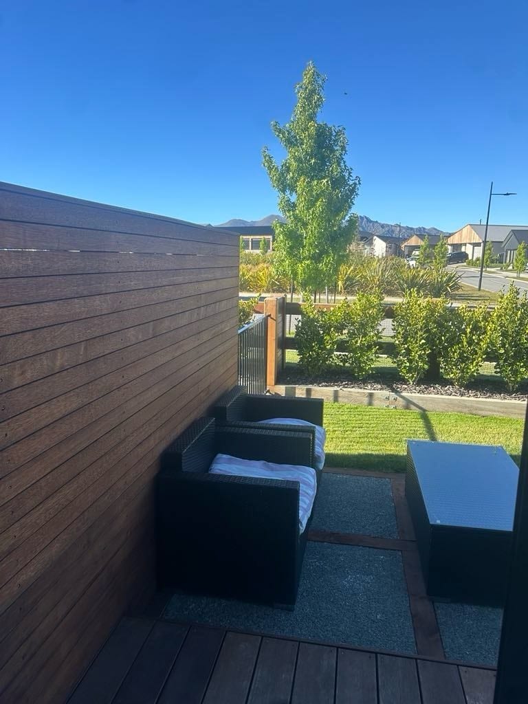 A private outdoor patio area is shown, featuring two dark wicker chairs with white cushions beside a matching coffee table. Lush greenery and a solitary tree border the space, offering a view of the surrounding neighborhood and distant mountains under a clear blue sky.