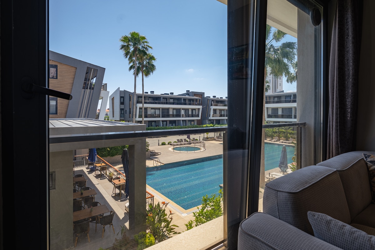 A spacious view is provided from the window, showcasing the outdoor pool surrounded by lounge chairs and palm trees. The modern architectural design of nearby buildings adds to the serene atmosphere. Natural light illuminates the interior space, creating a welcoming ambiance.