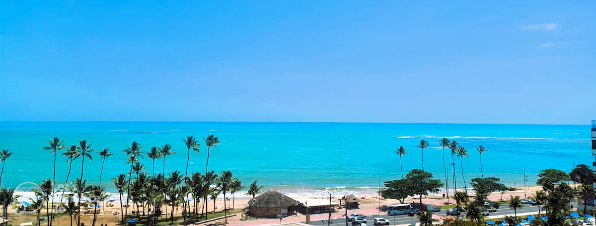 The image captures a serene beachfront view, featuring gentle waves lapping against the shore. Lush palm trees line the coastline, and a thatched-roof structure can be seen on the sand. The sky is clear, with vibrant shades of blue creating a peaceful backdrop.
