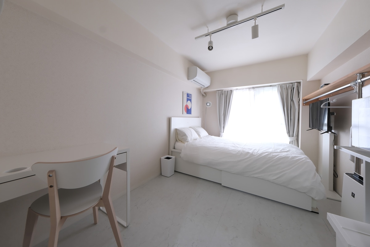 A bright and spacious room features a comfortable bed with white linens. A desk with a chair is set against one wall, while a TV is mounted opposite. Natural light filters through the window, illuminating the light-colored walls and floor.