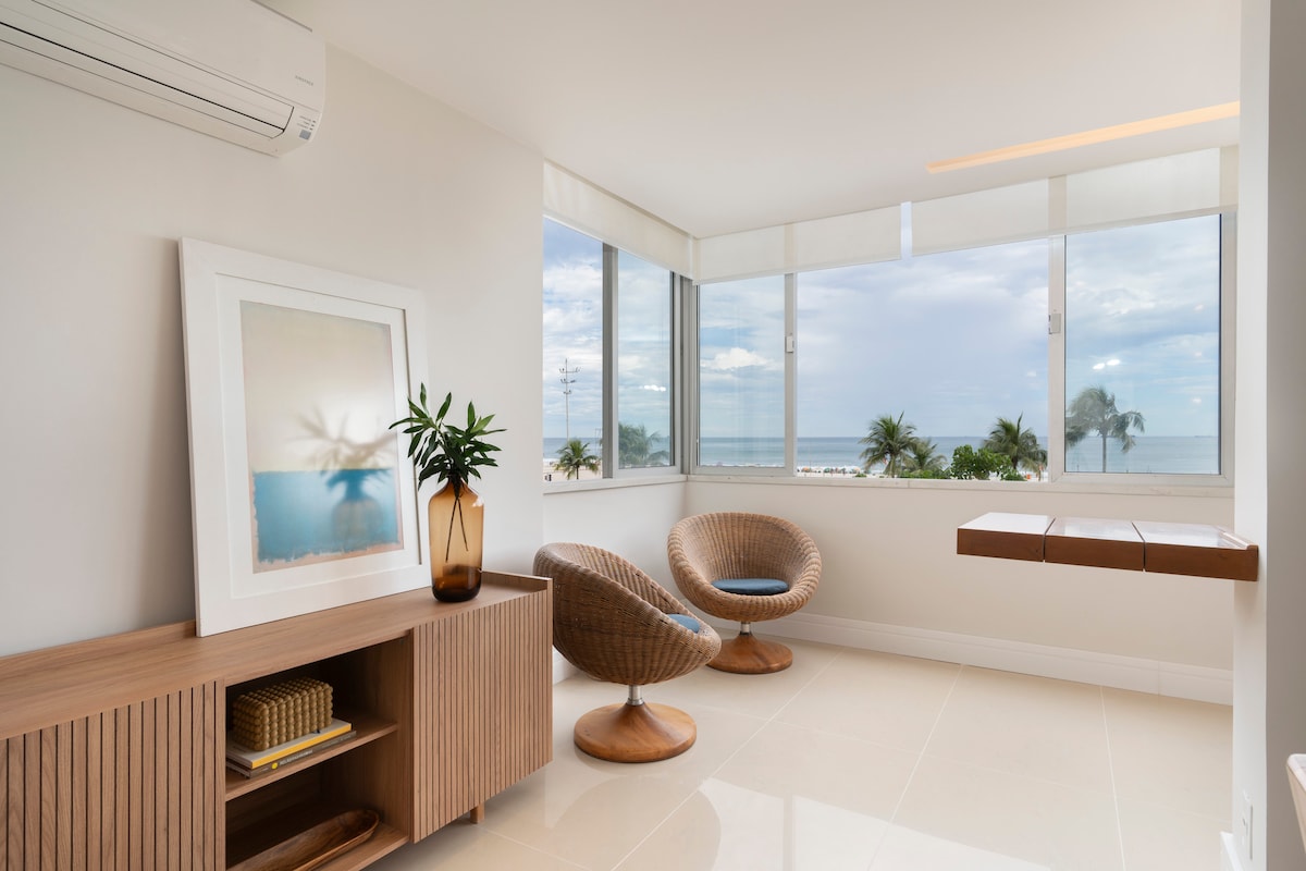 A sunlit corner of the apartment features large windows providing a view of the ocean and palm trees. Two rounded chairs are positioned near a sleek wooden table, complementing the minimalist decor. A framed artwork rests on a wooden console, enhancing the serene ambiance.