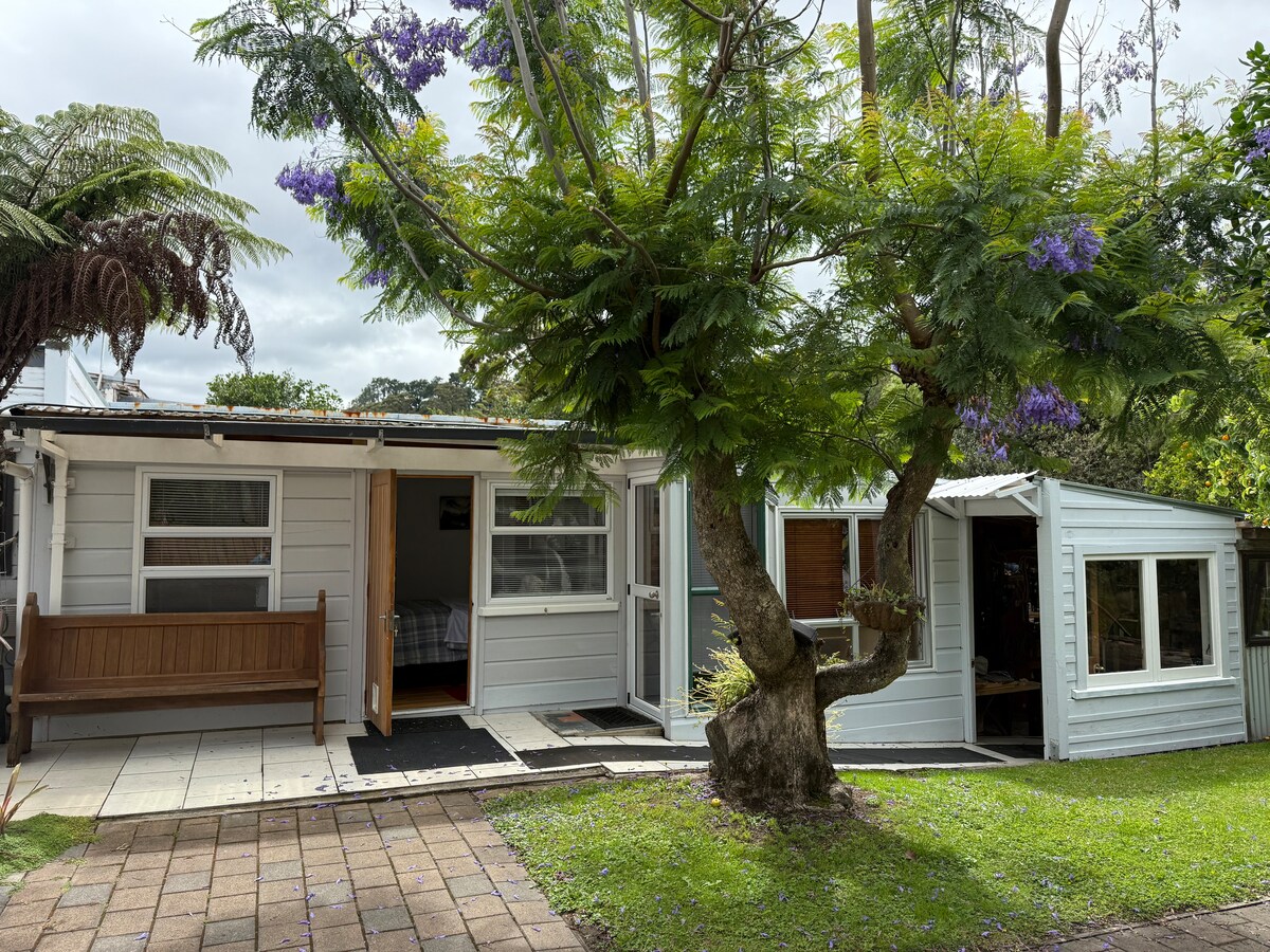 A charming exterior of a light-colored cottage is showcased, featuring a wide entrance and several windows. Lush greenery surrounds the structure, with a tree adorned with purple blooms contributing to the inviting landscape. A small patio area is visible, offering a space for relaxation.