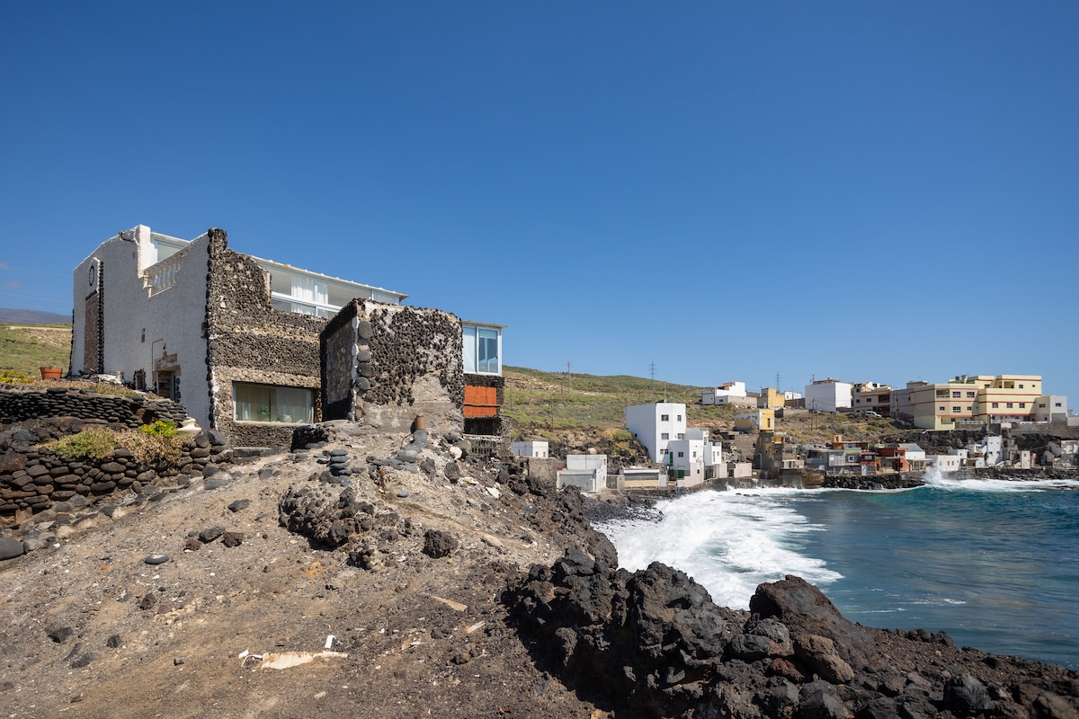 The apartment is positioned prominently on rocky terrain by the ocean, showcasing a mix of natural stone and modern architecture. Waves crash against the shoreline below, while a coastline of homes stretches into the background under a clear blue sky.