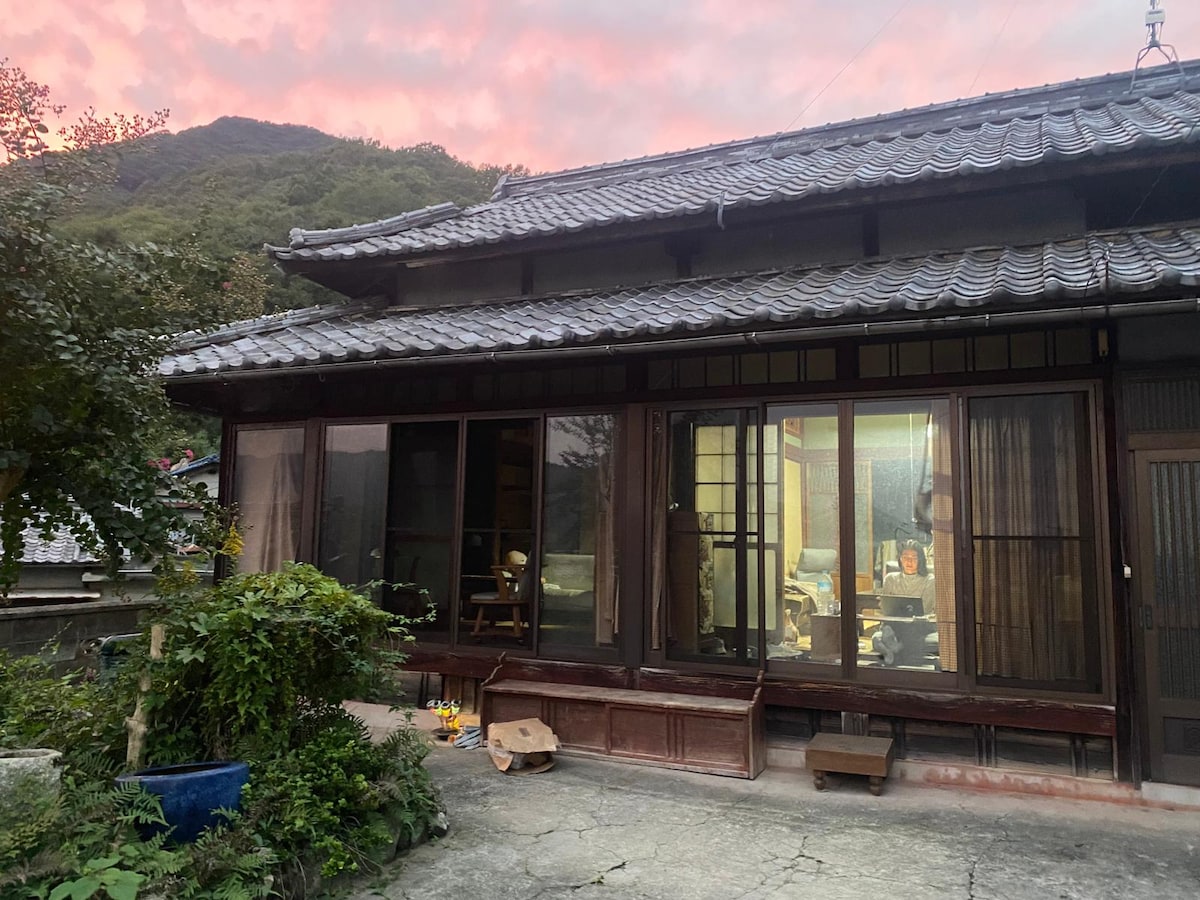 The exterior of a traditional-style house is depicted, showcasing a sloped tiled roof and large glass doors. Nearby greenery is visible, with various plants surrounding the entrance. A warm glow emanates from within, hinting at a welcoming interior as the sky transitions to shades of pink.