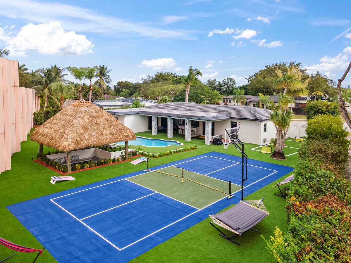 An outdoor area features a blue sports court with a pickleball net and a basketball hoop. A thatched-roof gazebo is visible nearby, accompanied by a swimming pool. Hammocks are set up on the lush green lawn, surrounded by palm trees and colorful landscaping.