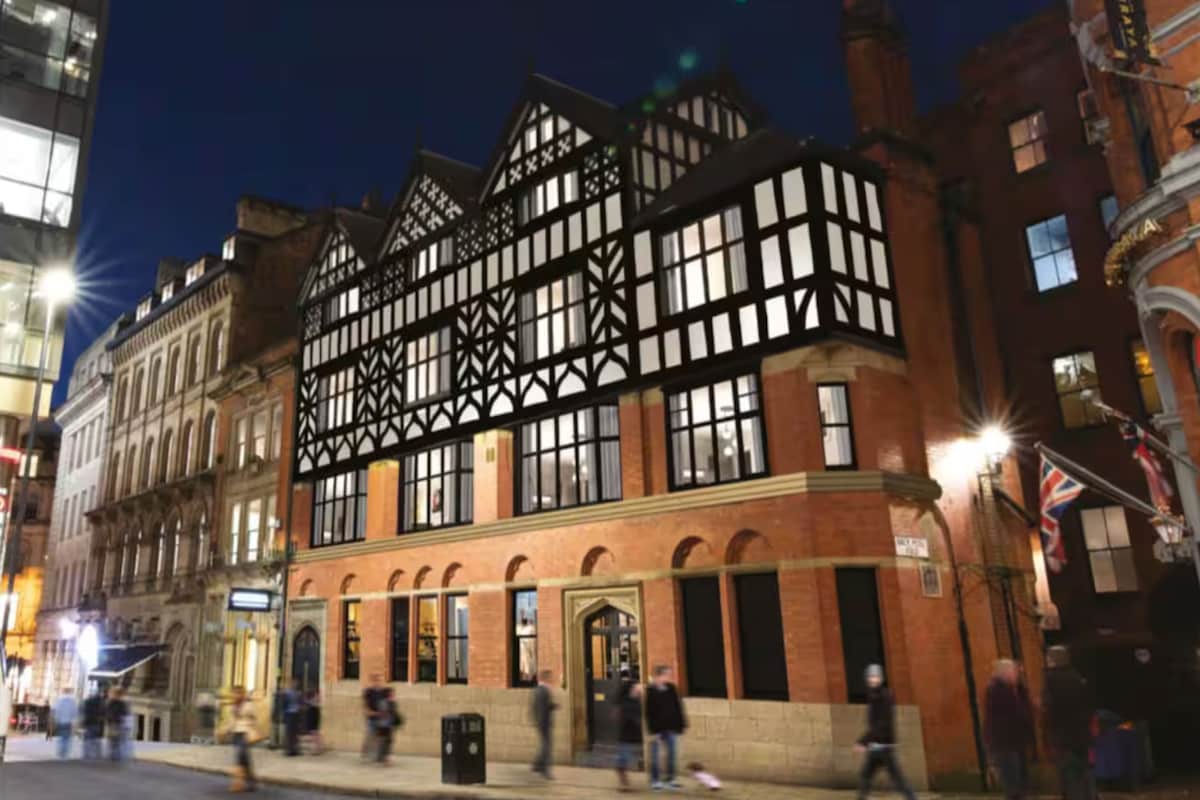 The building features a distinctive architectural style, showcasing a combination of brick and timber elements. The façade is illuminated at night, highlighting its unique character. Pedestrians are seen walking along the street, adding to the vibrant urban atmosphere.