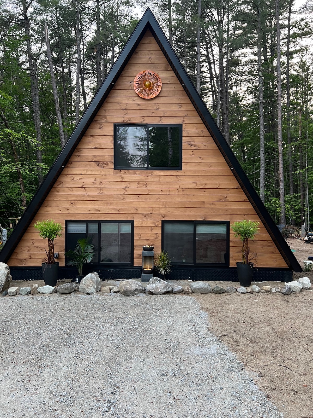 An A-frame structure is showcased, featuring a wooden exterior with large windows on the lower level. Flanking the entrance, black planters contain greenery. A sun-shaped decoration is positioned above the central window, while a gravel path leads up to the home amidst surrounding trees.