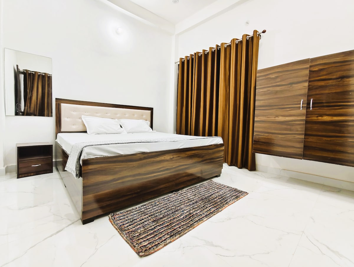 A spacious room features a polished wooden bed with a light-colored upholstered headboard. Dark curtains frame a window, complementing the room's neutral tones. A matching wooden wardrobe stands against one wall, while a nightstand rests beside the bed. A textured rug adds warmth to the floor.