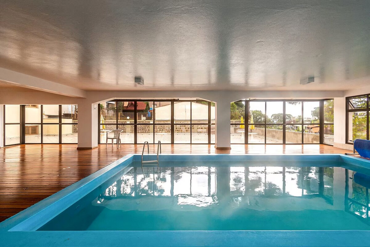 An indoor swimming pool is surrounded by large glass windows, offering views of the outdoors. The water's surface reflects the ceiling lights, while wooden flooring adds warmth to the space. A ladder is visible for ease of access into the pool.