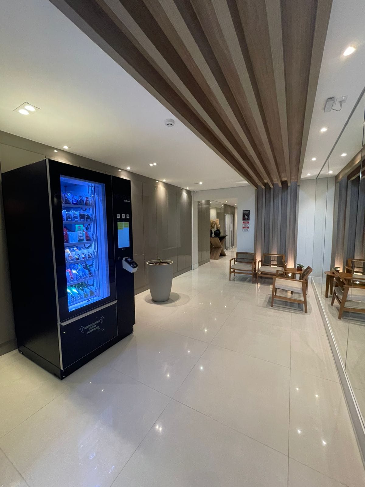 A modern hallway is illuminated by soft lighting, featuring a sleek vending machine in an eye-catching black finish. Adjacent to the machine, two minimalist wooden chairs are positioned, providing a casual seating area. The polished floor reflects the warm tones of the ceiling design.