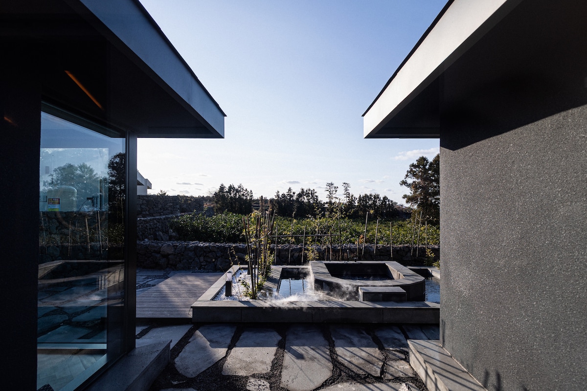 An outdoor space is featured between two modern structures, showcasing a tranquil setting with landscaped greenery and a jacuzzi. Large glass panels provide views of the natural surroundings, while stone pathways connect different areas, creating an open and inviting environment.