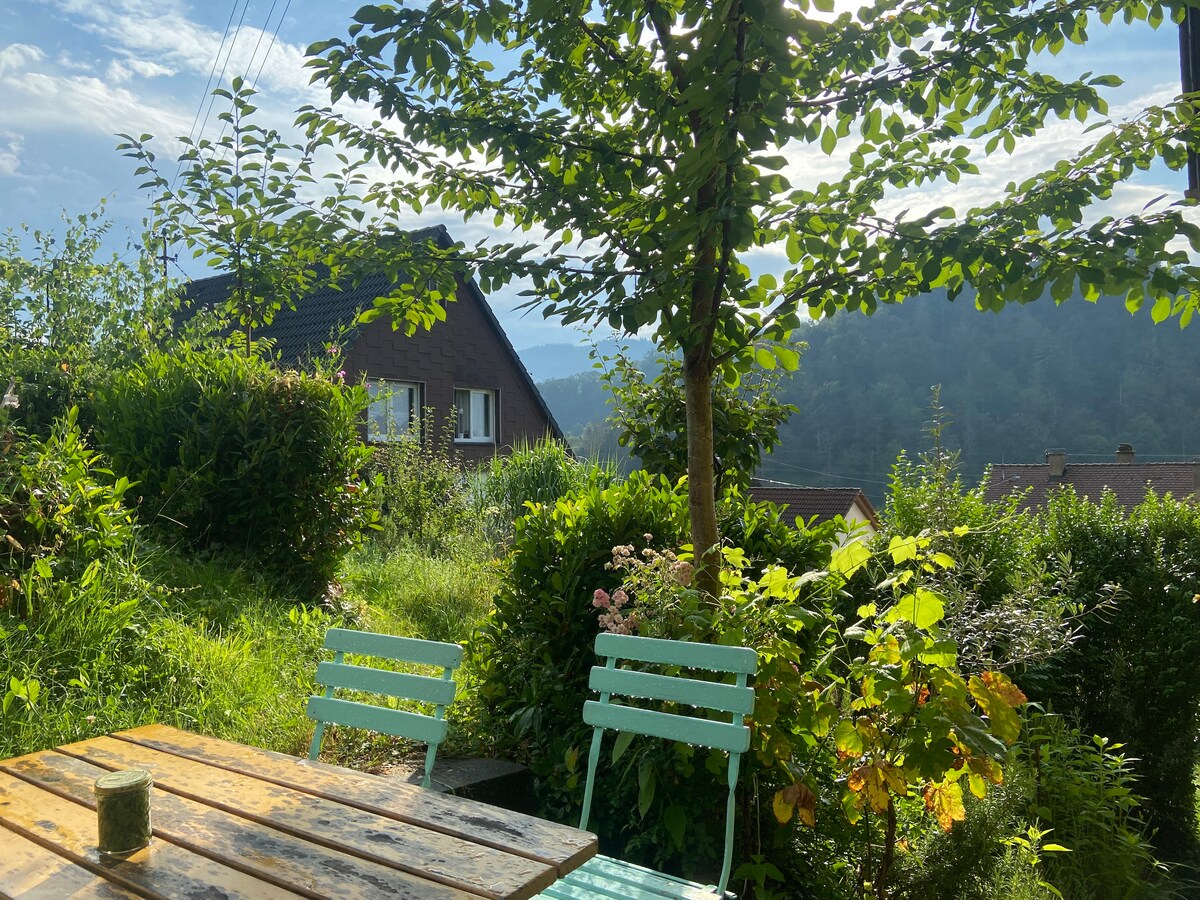 A garden area is depicted, featuring a wooden table with a green drink and two metal chairs. Lush greenery surrounds the space, including flowers and plants. In the background, a tree provides shade, with a view of homes and mountains under a clear sky.