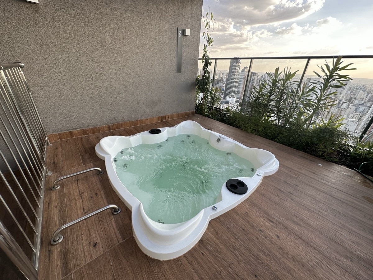 An outdoor hot tub is situated on a wooden deck, offering views of the city skyline. Surrounded by greenery, the tub features built-in seating with jets. The space is set against a backdrop of a gray wall and a railing, allowing for a serene atmosphere.