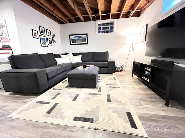 Sehemu ya ziada ya kuishi katika chumba kikubwa cha chini kilichokamilika chenye televisheni ya inchi 65, upau wa sauti, meza na viti. 