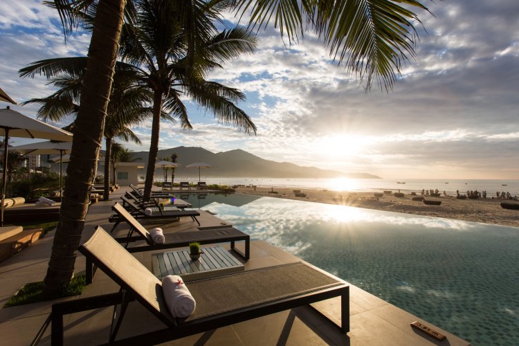 A serene beachfront scene is captured, showcasing comfortable lounge chairs positioned beside an inviting swimming pool. Palm trees gently sway in the breeze, while the sun sets over the ocean, casting a warm glow across the tranquil water and distant mountains.