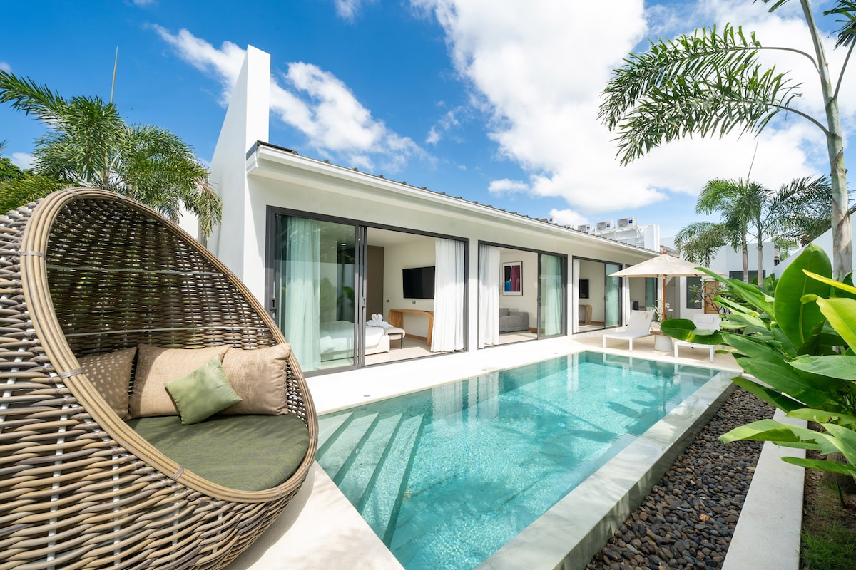 A modern villa is seen with large sliding glass doors leading to a private pool. A round woven chair is placed next to the pool, surrounded by lush greenery and palm trees. The clear blue sky enhances the serene atmosphere of the outdoor space.