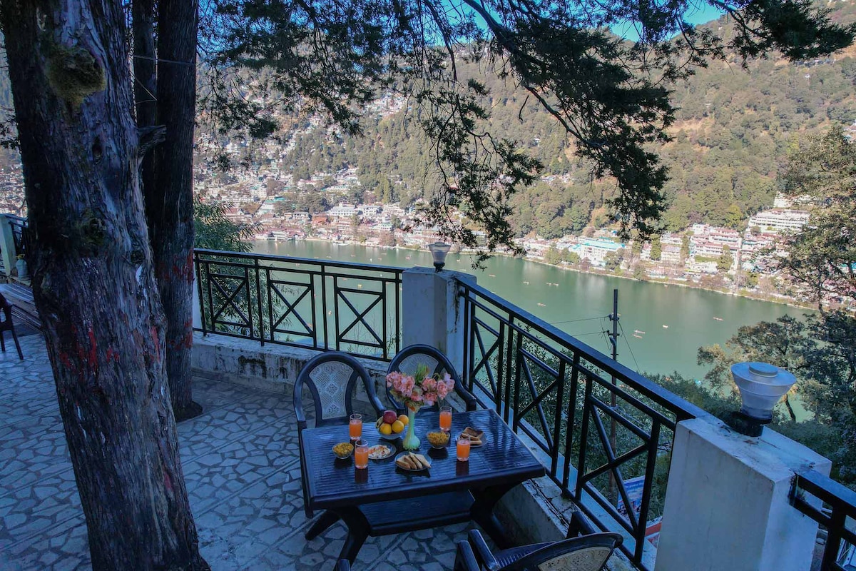 A dining table is set on a balcony overlooking Naini Lake, surrounded by lush greenery. The table is adorned with fruit, flowers, and refreshing drinks. Scenic hills and colorful boats on the water create a serene backdrop, inviting relaxation and enjoyment.