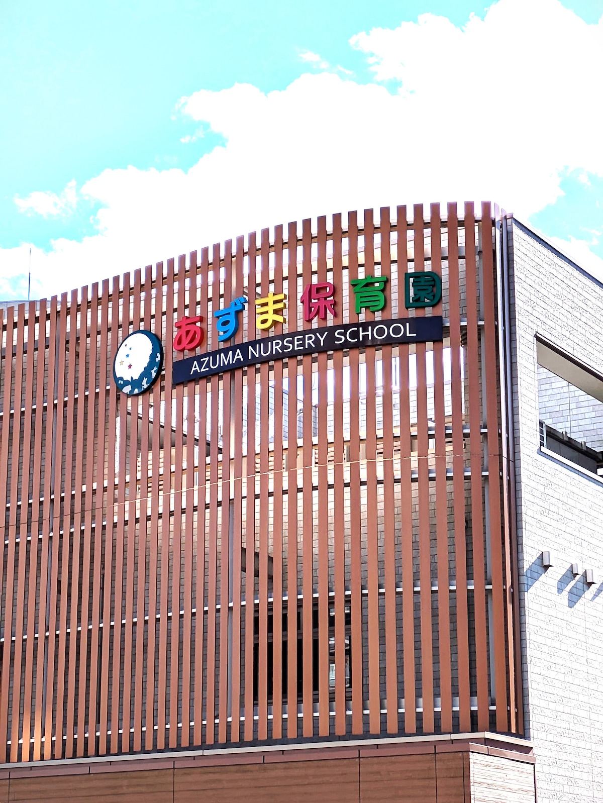 A modern nursery school building with a wooden slatted facade features a colorful sign displaying the name 'Azuma Nursery School.' A clock is mounted near the sign, complementing the clean architectural lines under a clear blue sky.
