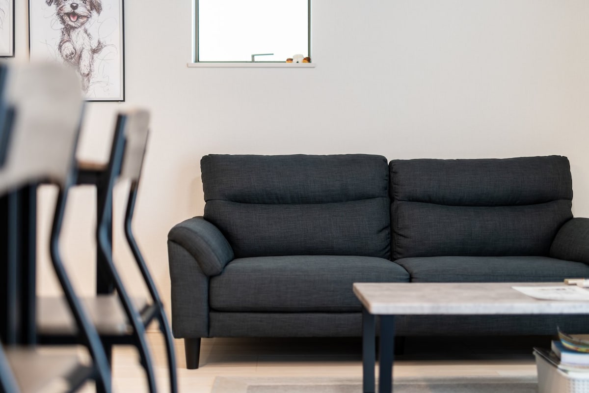 A dark gray sofa is positioned against a light-colored wall, with a small window situated above it, providing natural light. Artistic frames are visible on the wall, adding character to the space, while a wooden coffee table is placed in front of the sofa.