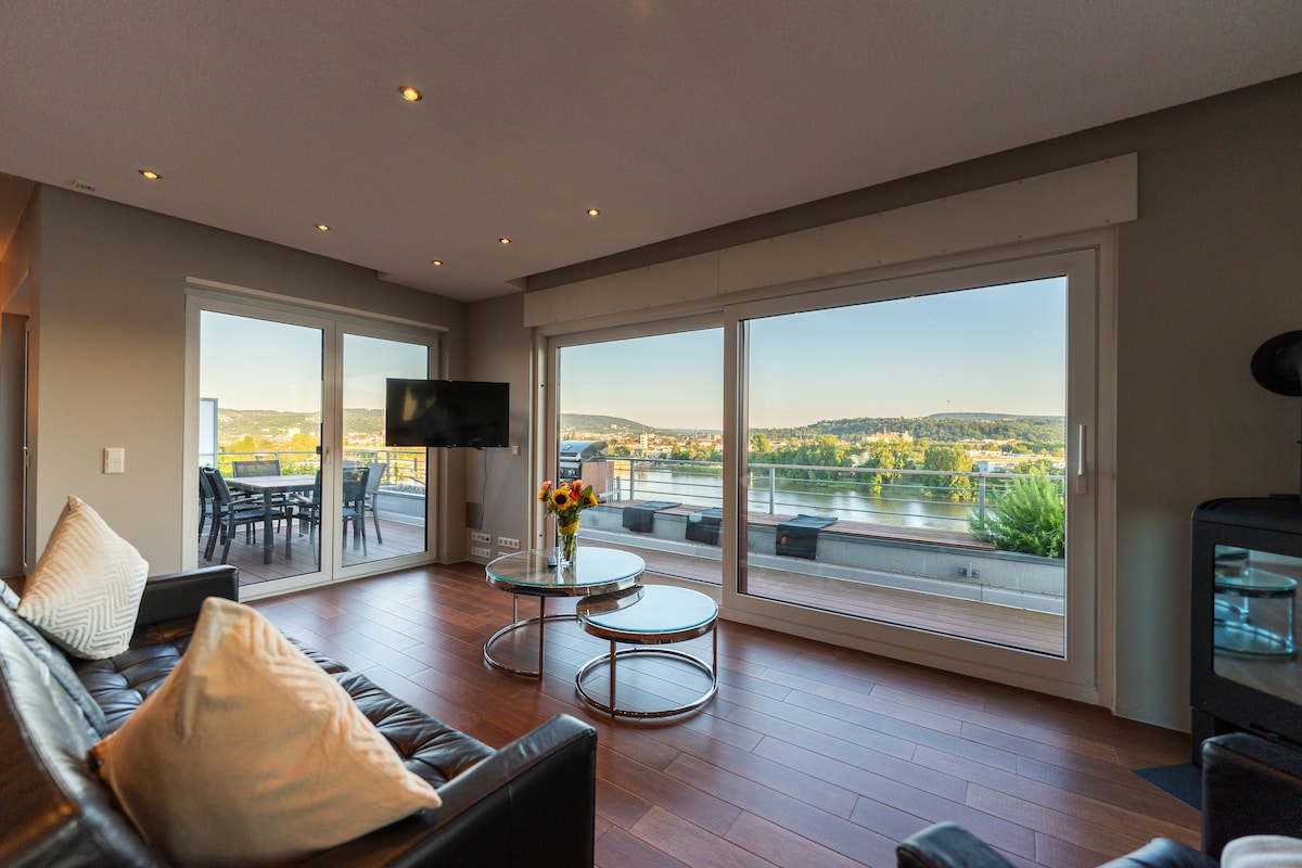A spacious living area features large glass windows offering views of the Mosel River. A comfortable black leather sofa is arranged around a glass coffee table, with a television mounted on the wall. Natural light fills the room, enhancing the inviting ambiance.