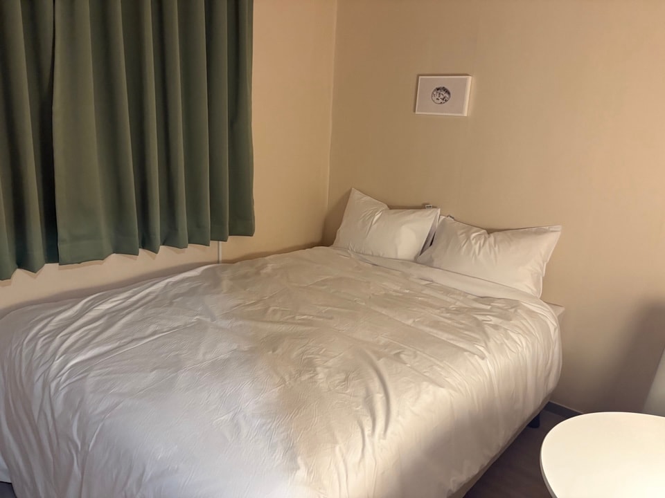 A cozy sleeping area features a spacious bed with white bedding, flanked by two white pillows. A small round table is positioned nearby, and green curtains partially cover a window, allowing for a serene atmosphere.